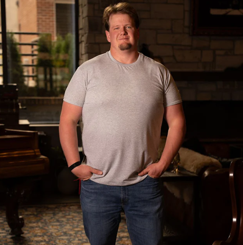Man wearing a light gray heather t-shirt in size 3XL and blue jeans, standing in a rustic, upscale living room.