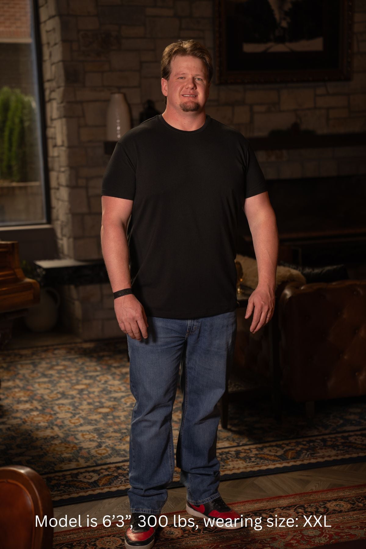6'3", 300 pound man wearing a black XXL t-shirt and blue jeans standing in a room with a brick wall and couch.