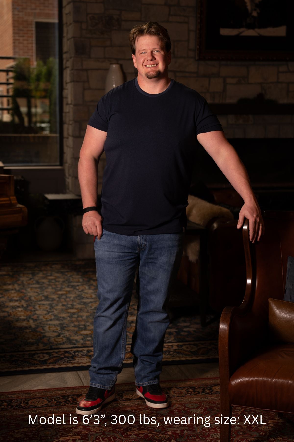 6'3", 300 pound man wearing a black XXL t-shirt and blue jeans standing in a room with a stone wall and brown leather chair.