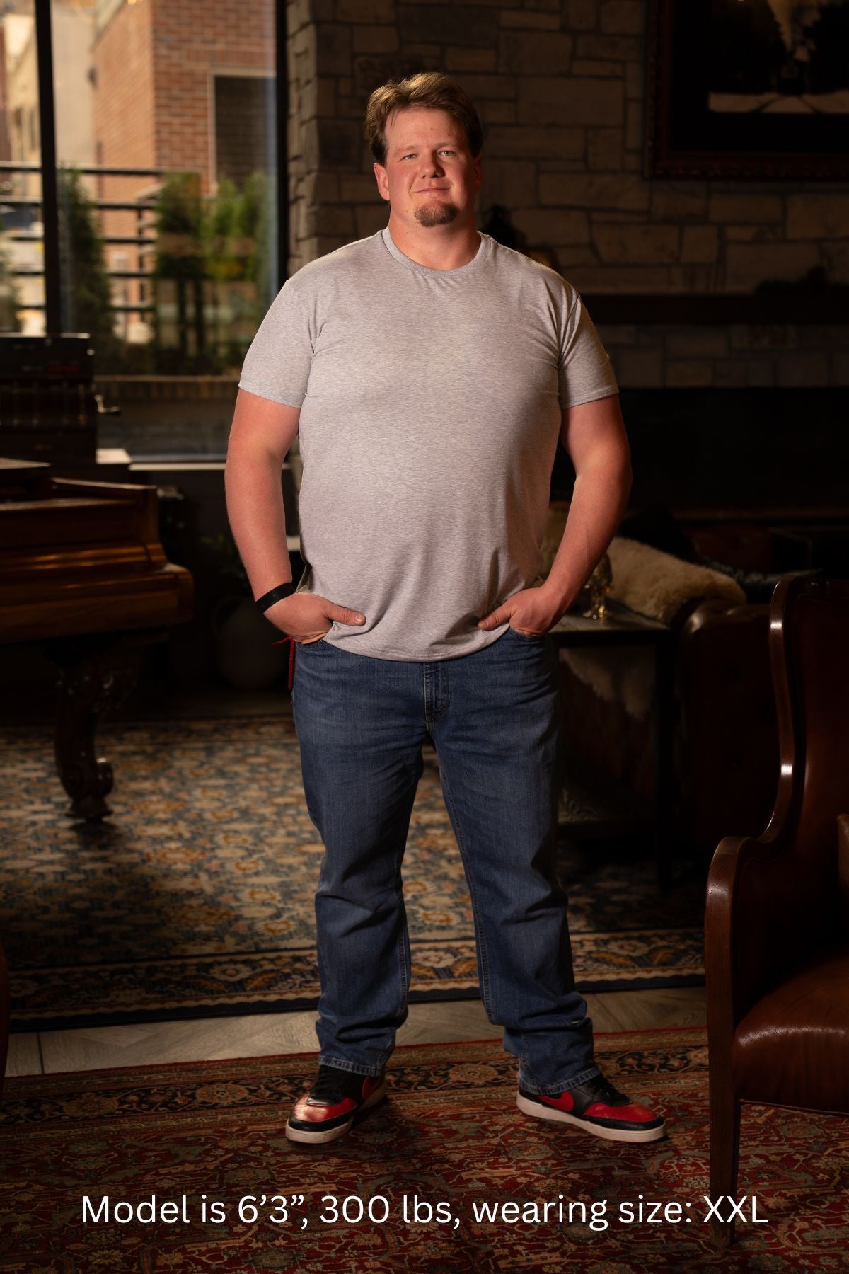 6'3", 300 pound man wearing a XXL gray t-shirt and blue jeans standing in a room with a brick wall and wooden furniture.