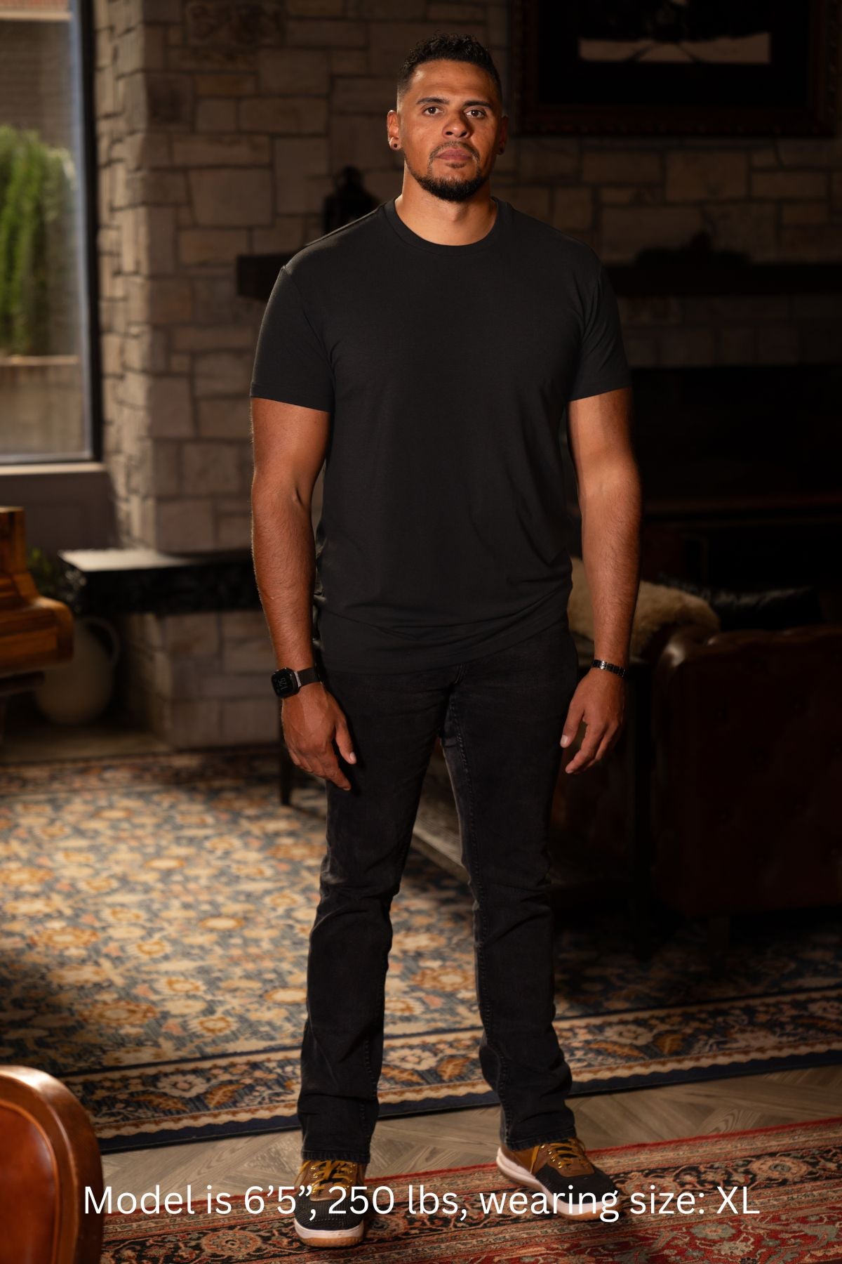 6'5", 250 pound man wearing a black XL t-shirt and pants standing in a room with a stone wall and rug.